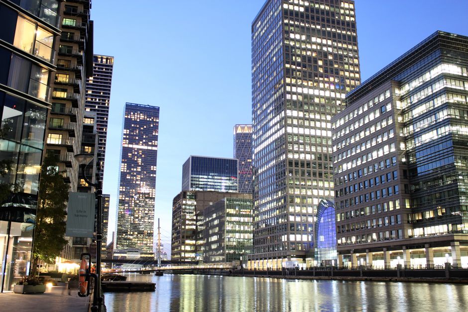 Nighttime view of Canary Wharf E14 with modern commercial office buildings featuring glass and steel facades, illuminated windows reflecting on the waterway, and a clear sky above. The scene includes 