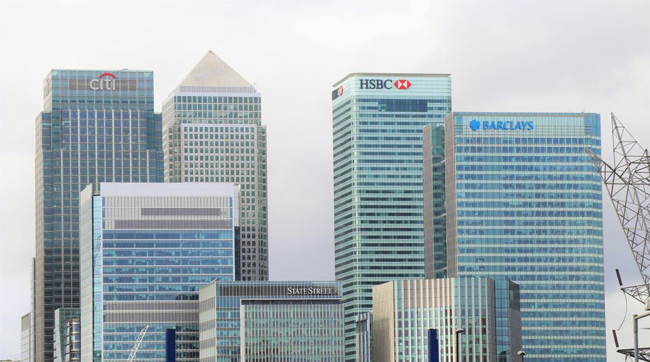 Nighttime view of Canary Wharf E14 with modern commercial office buildings featuring glass and steel facades, illuminated windows reflecting on the waterway, and a clear sky above. The scene includes 
