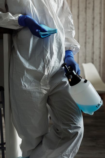 A professional cleaning technician from carpetclean.london wearing a white protective suit and blue gloves, holding a spray bottle filled with cleaning solution in one hand and a blue microfiber cloth