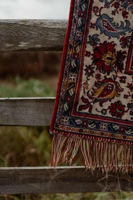 A detailed close-up of a traditional Persian-style rug hanging over a weathered wooden fence outdoors. The rug features intricate floral patterns in shades of red, blue, yellow, and beige, with fringe