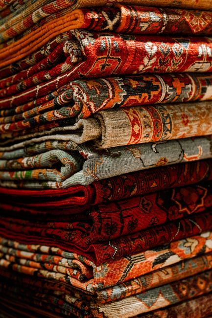 A detailed close-up of a traditional Persian-style rug hanging over a weathered wooden fence outdoors. The rug features intricate floral patterns in shades of red, blue, yellow, and beige, with fringe